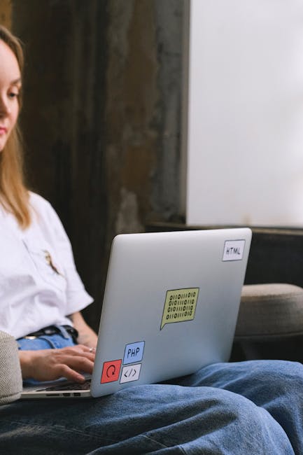 A person with long brown hair wearing a white shirt sits on a sofa with their legs crossed, working on a silver laptop that features colorful stickers, including a yellow one with binary code, a blue